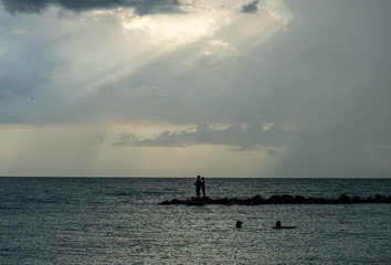 beach in Florida on the Gulf of Mexico with a cloudy sky with light shining through and rain in the distance.  A lone fisherman stands on the jetty