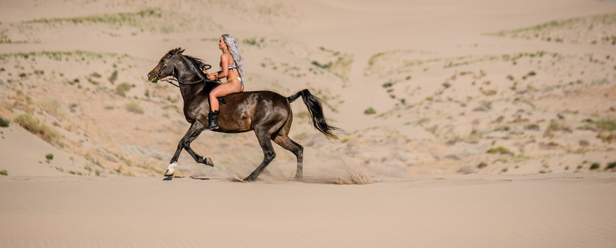 blonde woman in bikini on dark brown Arabian horse racing through the sand
