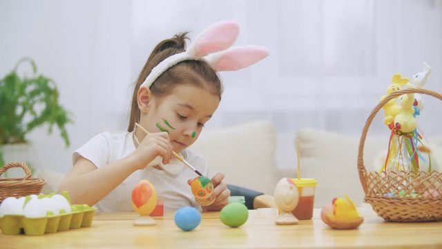 Nice, cute girl is having fun painting an Easter egg. Adorable girl is colorizing and Ester egg . Girl with beauty spots at her face and is smiling gently with cat's whisker, sitting at the wooden