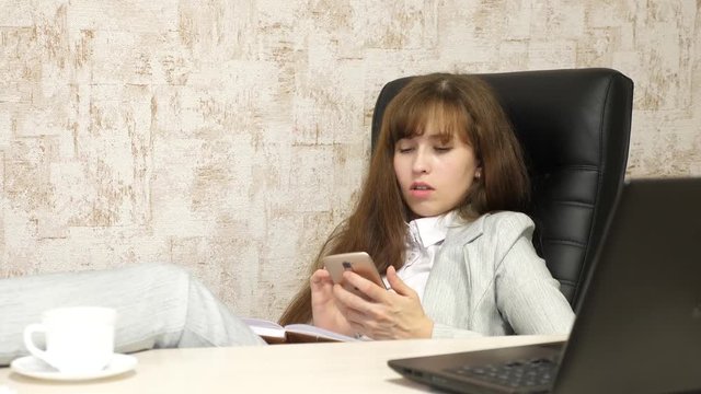 woman in office resting in workplace. beautiful businesswoman with phone sits in a chair with bare feet on the table resting. a girl at work in the office writes letter on her smartphone phone.