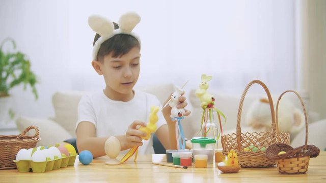 Young adorable boy is sitting at the table full of Easter decorations and is playing with Easter bunnies in his hands. Bunnies' discussion. Who would paint an egg Bunny chooses green colour. Bunny