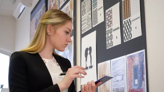 Attractive young girl in stylish clothes by mood board. White caucasian girl make notes on clipboard by pen. Young woman write and draw scetch near mood creative board