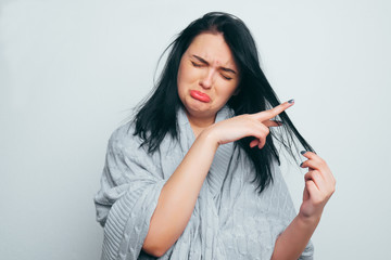 Fototapeta premium Upset Young beautiful girl in a gray robe, folding her fingers in the form of scissors cuts off a strand of hair. Split ends, it's time to get a haircut on a gray-white studio background