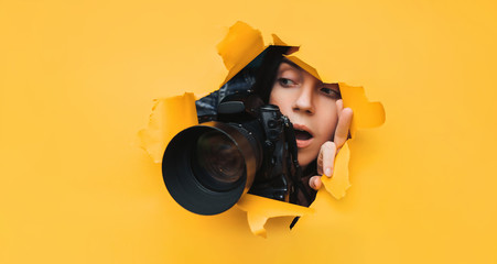 A paparazzi girl with a reflex camera looks out from cover and looks at what is happening with her mouth open. Yellow paper, torn hole. Tabloid press. In search of the plot for photo stocks. - obrazy, fototapety, plakaty