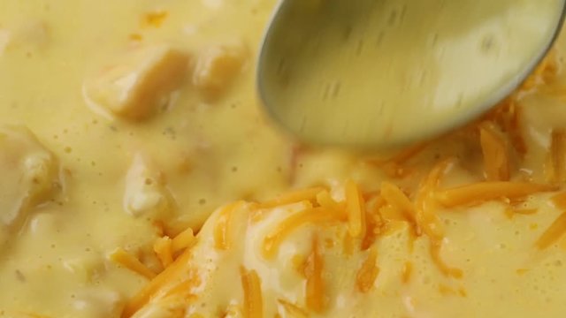 Very close video of stirring shredded cheddar cheese into chicken corn chowder with a spoon while cooking illuminated with natural lighting.