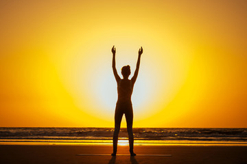 Fototapeta premium Silhouette of young woman in a stylish suit for yogi jumpsuit doing yoga on the beach in pose copy space