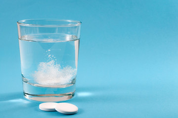 Recovering from a hangover and nursing a headache with aspirin concept with effervescent drink tablet dissolving in water with two tablets outside the glass isolated on blue background with copy space