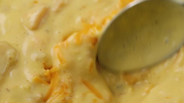 Close video of stirring shredded cheddar cheese into hot chicken corn chowder with a spoon illuminated with natural lighting.
