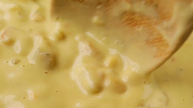 Very close video of stirring hot chicken corn chowder with a wood spoon while heating illuminated with natural lighting.