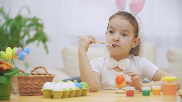 Little playful girl, wearing bunny ears on her head chose a red colour to paint an egg. Girl has painted an heart on it. Girl is cleaning her nose and is putting a paint-brush near to her mouth and