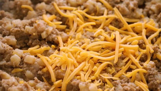 Close video of adding extra cheddar cheese to sausage hash in a pan while cooking illuminated with natural lighting.