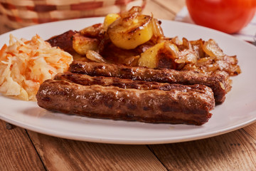 Serbian cevapi, cevapcici, Balkan minced meat kebab on a white plate with marinated cabbage, fried potatoes and fried onions next to bread and tomat on a wooden background