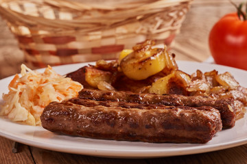Serbian cevapi, cevapcici, Balkan minced meat kebab on a white plate with marinated cabbage, fried potatoes and fried onions next to bread and tomat on a wooden background