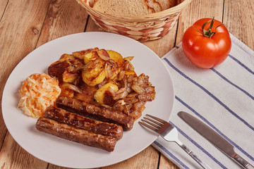 Serbian cevapi, cevapcici, Balkan minced meat kebab on a white plate with marinated cabbage, fried potatoes and fried onions next to bread, tomat and cutlery on a wooden background