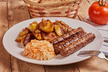 Serbian cevapi, cevapcici, Balkan minced meat kebab on a white plate with marinated cabbage, fried potatoes and fried onions next to bread, tomat and cutlery on a wooden background