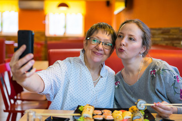 Business grandmother and young girl sit in a restaurant and take a selfie on the phone. We ordered sushi and rolls for dinner.