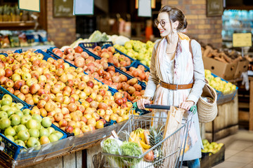 Young woman buying food, standing with shopping cart full of products in the supermarket with fruits on the background