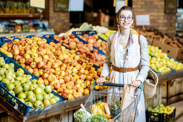 Young woman buying food, standing with shopping cart full of products in the supermarket with fruits on the background