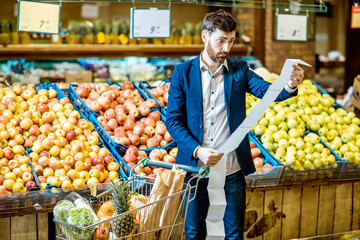 Portrait of an elegant man with shocked emotions holding very long shopping list while buying food in the supermarket