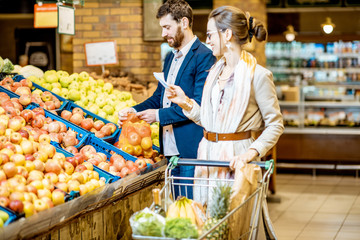 Young and happy couple buying fresh fruits standing together with shopping list and cart in the...