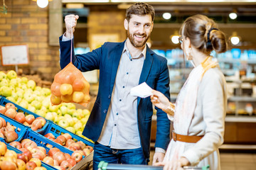 Young and happy couple buying fresh fruits standing together with shopping list and cart in the...