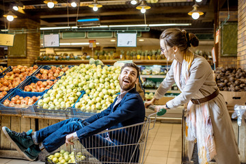 Man and woman having fun while riding with shopping cart in the supermarket with fruits and...