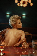 Young beautiful blonde girl in a luxurious golden sexy dress sitting at the bar.