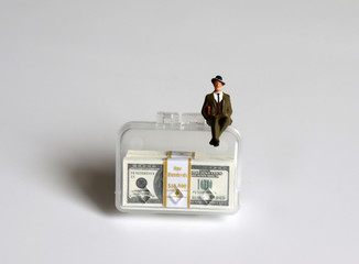 A miniature businessman sitting on a bag containing a hundred-dollar bills.