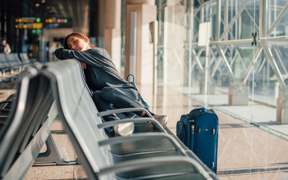 Young tired woman sleeping alone in empty airport with her hand luggage, waiting flight - transportation, low cost traveling, delayed or cancelled flight concept