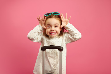 Cute little girl in sunglasses makes funny crazy face, big brown eyes, and raised arms stands near...