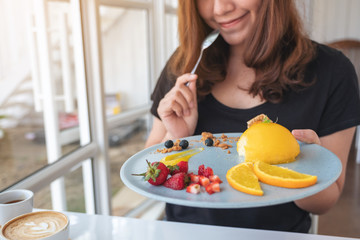 A beautiful asian woman holding a plate of orange cake with mixed fruit and a spoon in cafe