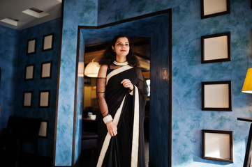 Young indian woman wear at elegant black saree posed on restaurant against wall with frames.