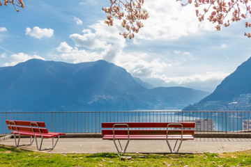 The Magnolia branches and the two empty red benches at the hill in Lugano, view at the Lake Lugano and Alps mountains in Ticino canton of Switzerland