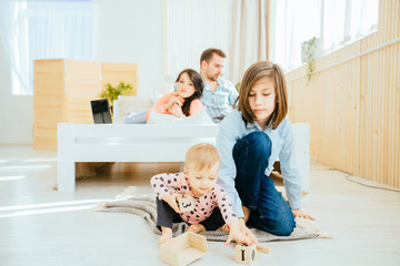 Brother and little sister playing together sits on the floor, while their freelancers parents working with laptop lying on bed on background. Happy family together relations concept.