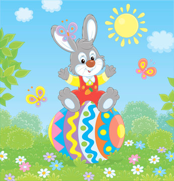 Little Easter Bunny sitting on a colorfully decorated big egg among flowers and flittering butterflies on a green lawn on a sunny spring day, vector illustration in a cartoon style