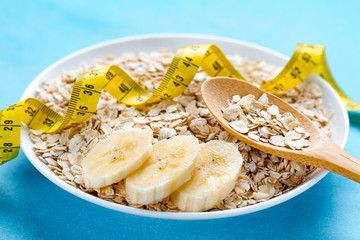 Diet concept. Oatmeal bowl and spoon with fresh ripe banana and yellow measuring tape on blue background. Oat flakes for healthy and fiber breakfast, slimming food