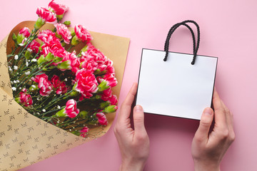 Obraz premium Bouquet of fresh carnations and a paper white gift bag on pink background. Receiving gifts and flowers from loved ones