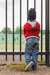 Kid boy stands by the fence and looks into the distance. Metal fence around the stadium or school. The boy looks at the guys playing on the field.