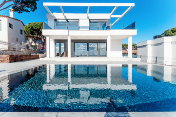 Luxury modern white house with large windows overlooking a Mediterian landscaped garden with palm trees and  blue swimming pool. High tech style villa. Vacation home or hotel. Modern loft design.ees a