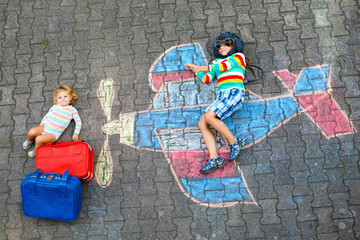 Two little children, kid boy and toddler girl having fun with with airplane picture drawing with colorful chalks on asphalt. Siblings painting with chalk and going on vacations.