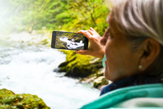 Nice attractive elderly mature woman with shiny grey hair taking photos and selfies outdoor in a Vintgar river canyon gorge on a beautiful sunny autumn day