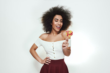 Sweet and beautiful. Cute and young afro american woman with curly hair looking at colorful lollipop in hand and smiling, keeping one hand on hip