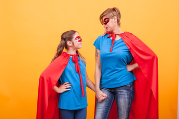 Mother and daughter dressed like superheros holding hands and looking to each other over yellow background.