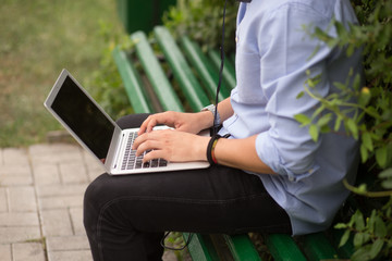 Fototapeta premium Cropped image of young man sitting at on the bench in the park, using a laptop and listening a music. Education concept, leisure.