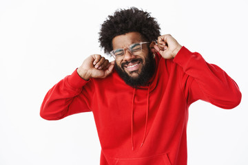 African american bearded guy in red hoodie grimacing, close eyes intense and clenching teeth as preparing to loud noise or sound of siren, closing ears not hear annoying bang over gray wall