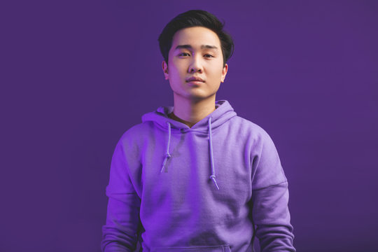 Portrait Of A Handsome Young Chinese College Student, Dressed In Casual Wear. Not Emotional Guy Looking At Camera With Serious Face, Isolated At Studio Against Puple Wall