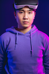 Half-size studio portrait of Asian cyberspace player with his vr glasses taken off , mounted on head, looking at camera isolated in casual clothes in dark blue background with copyspace
