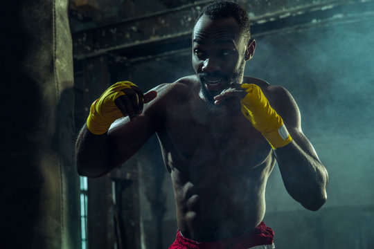 Hands of boxer over dark gym background. Strength, attack and motion concept. Fit african american model in movement. Naked muscular athlete. Sporty man during boxing.
