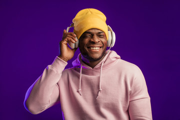 Joyful happy hipster man in white hoodie and yellow hat listening music with headphones, isolated at purple studio background. Hip hop style, positive emotions, face expression, dancing concept