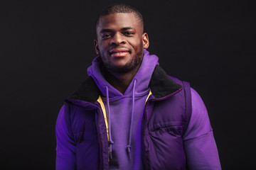 Positive smiling african man posing in sports fashion outdoor outfit on black background